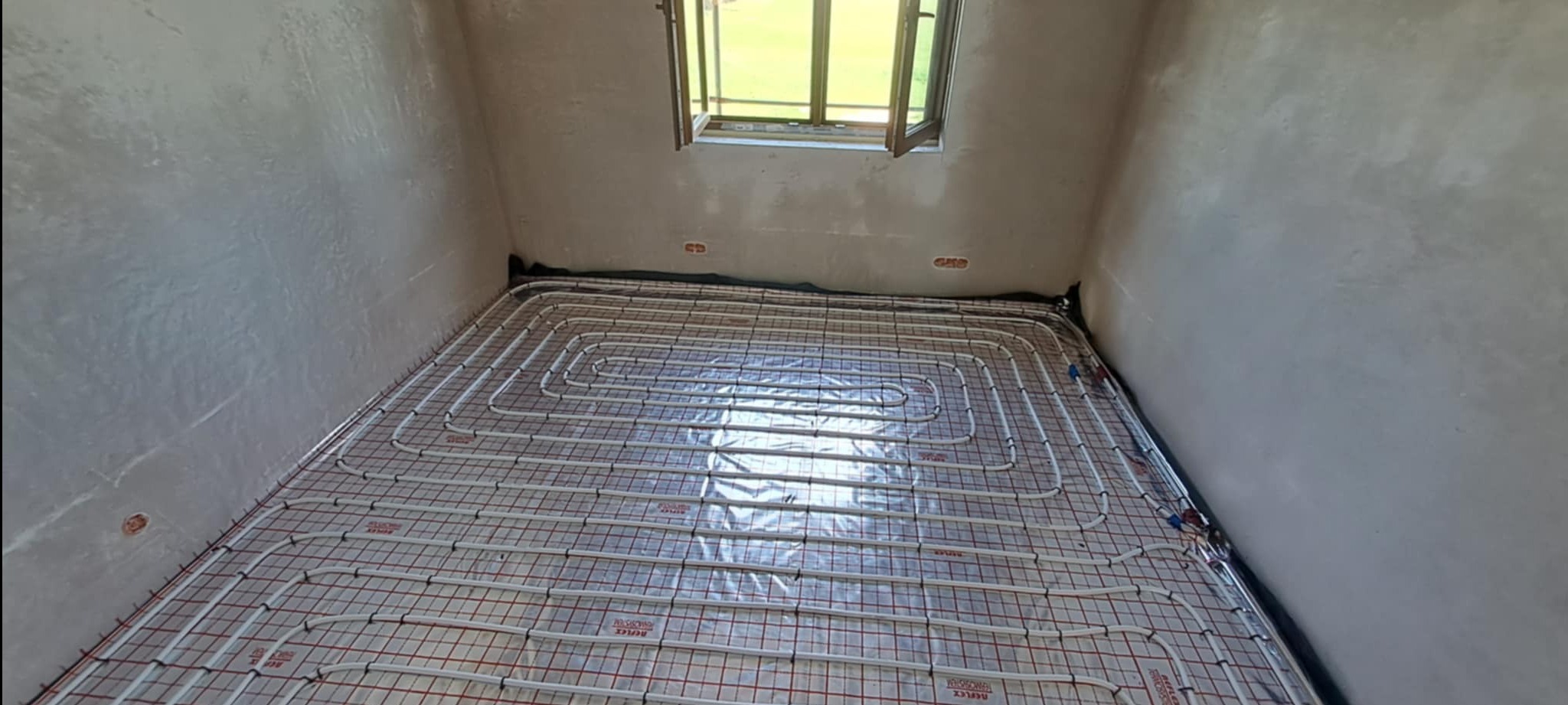 Widok z góry na podłogę z rozłożonym systemem ogrzewania podłogowego, rury ułożone w pętle na folii izolacyjnej z czerwoną siatką, surowe ściany i okno w tle.