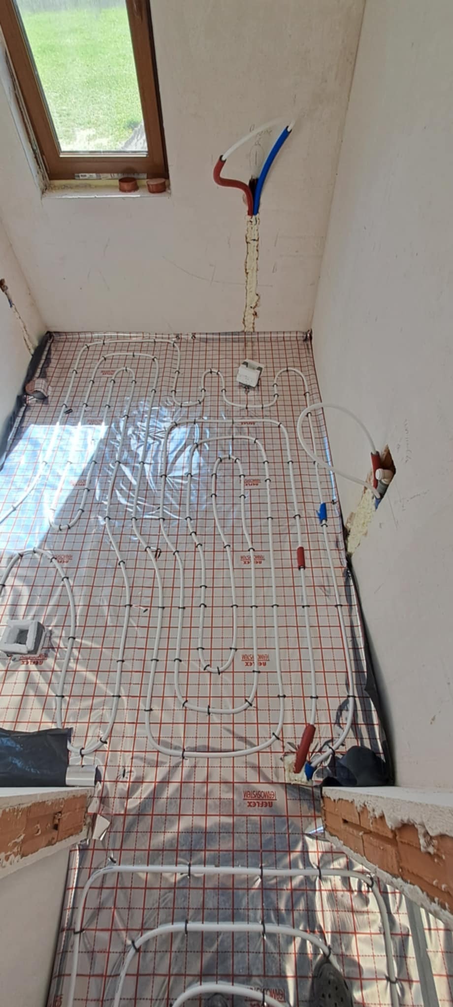 Podłogowe ogrzewanie wodne w trakcie montażu w Kowali, rury ułożone na siatce z folią odblaskową, widoczne przyłącza do instalacji.