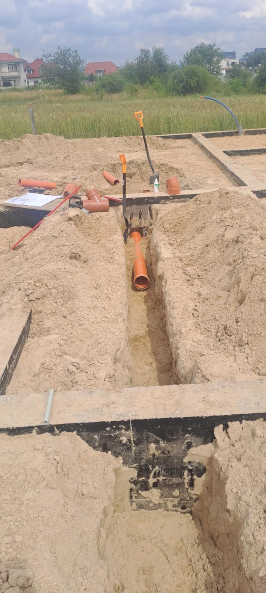 Pomarańczowe rury kanalizacyjne ułożone w wykopie w piasku, widoczne narzędzia, poziomica i dokumenty na obrzeżach wykopu, teren budowy w tle.