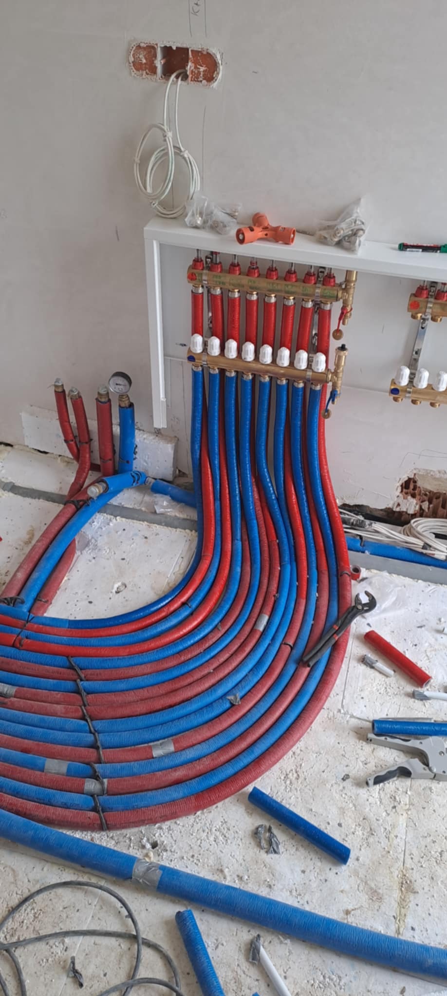 Rozdzielacz ogrzewania podłogowego z rurami PEX w kolorze niebieskim i czerwonym, manometr, narzędzia hydrauliczne i fragmenty rur na podłodze w trakcie montażu w surowym wnętrzu.