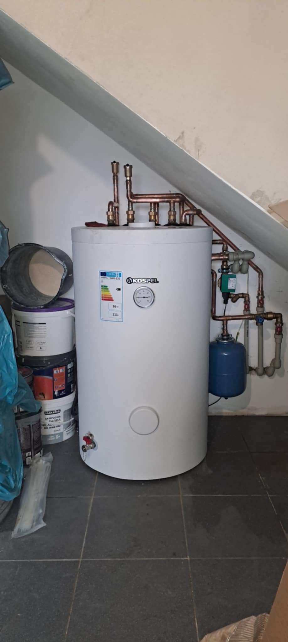 Biały bojler marki Kospel z miedzianymi rurami, manometrem i etykietą energetyczną, stojący na ciemnej podłodze z płytek, obok puszki z farbą i wiadro. Widoczne ukośne ściany poddasza.