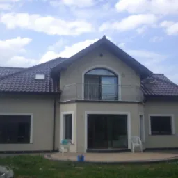 Dwupiętrowy dom z beżową elewacją i brązowym dachem, widoczny z ogrodu w słoneczny dzień. Balkon z metalową balustradą nad wejściem. Widoczne okna z białymi ramami.