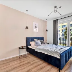 Sypialnia w Łodzi z drewnianą podłogą, granatowym łóżkiem z pikowanym zagłówkiem, czarnymi lampami wiszącymi i balkonem z widokiem na blok.