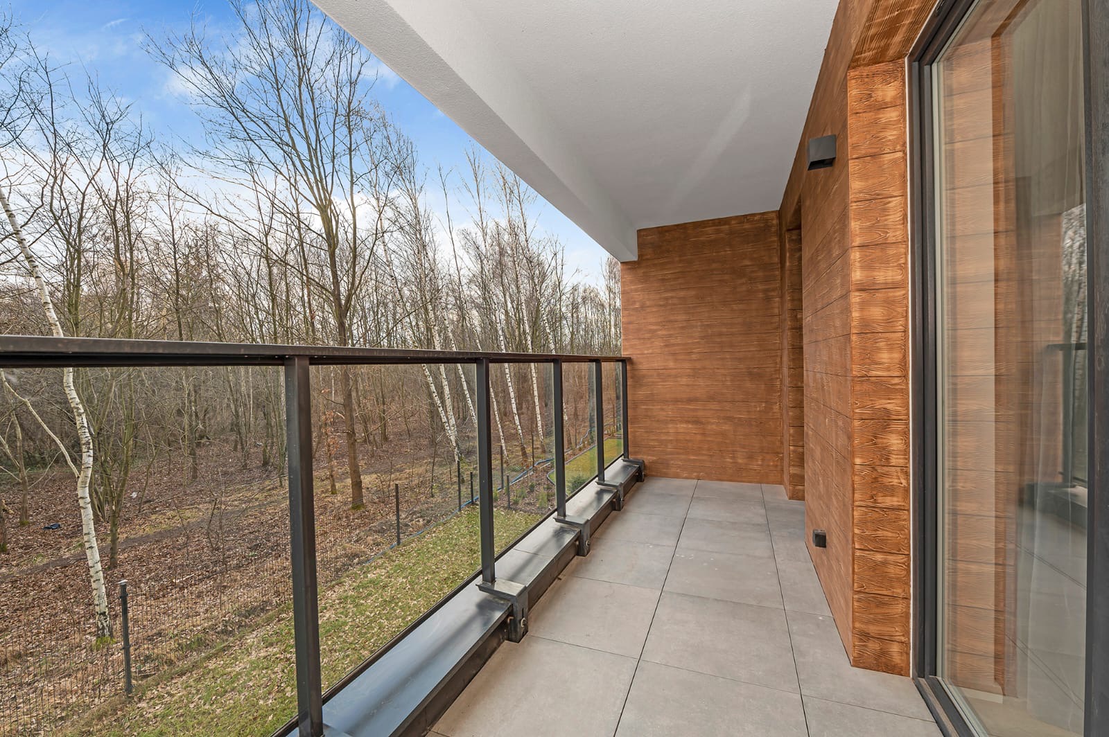 Nowoczesny balkon z szarą płytką podłogową, szklaną balustradą z czarnymi elementami i drewnopodobną okładziną ściany, widok na las.