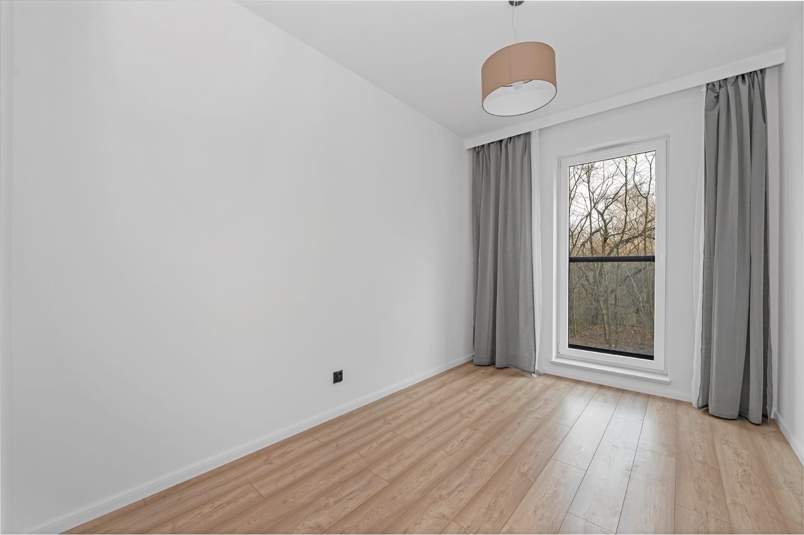 Pusty pokój z panelami podłogowymi imitującymi drewno, szare zasłony przy oknie z widokiem na drzewa bez liści, beżowa lampa sufitowa i czarny włącznik na białej ścianie.