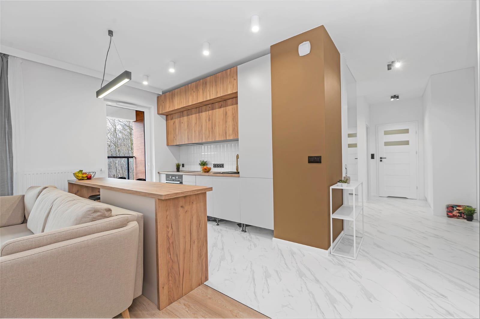 Nowoczesna kuchnia z drewnianymi szafkami górnymi, białymi szafkami dolnymi i marmurową podłogą, widok z salonu z beżową sofą i drewnianym blatem oddzielającym przestrzeń.