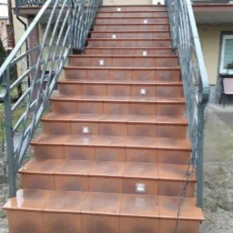 Zewnętrzne schody wejściowe wyłożone brązowymi płytkami z wbudowanym oświetleniem punktowym, balustrady z metaloplastyki, dekoracja w kształcie renifera w tle.