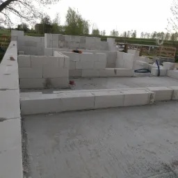 Wznoszenie ścian parteru z białych bloczków betonu komórkowego na wylewce fundamentowej, widoczny mur z wyżłobieniami, pomarańczowy pędzel na ścianie, w tle zieleń drzew i pastwisk.