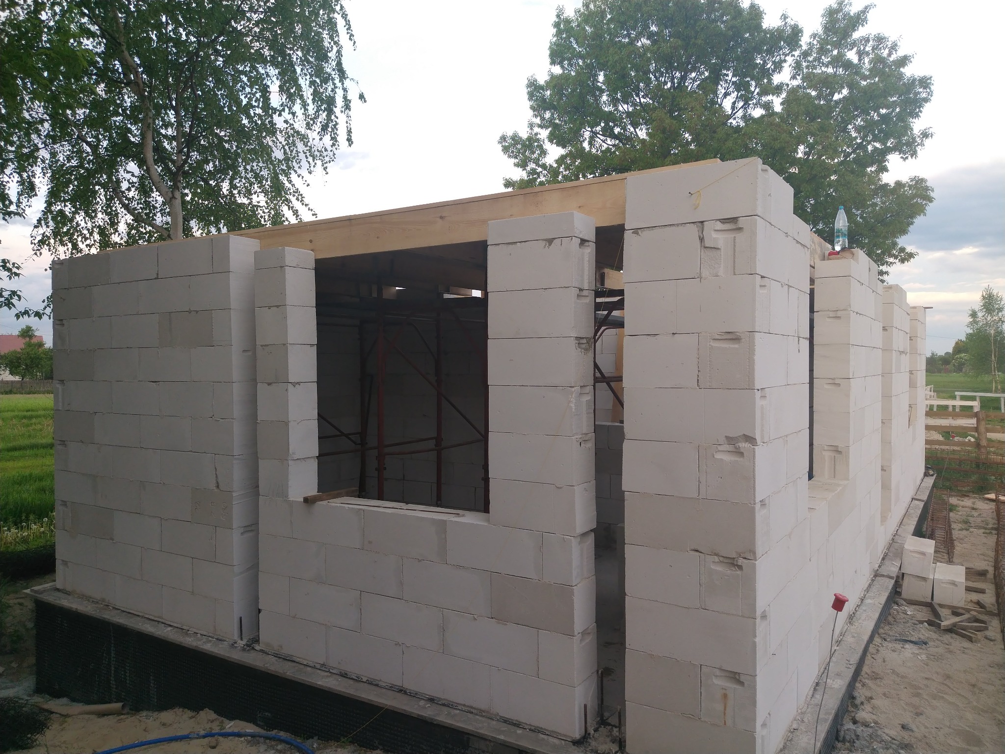 Surowy stan budowy domu z białych bloczków, widoczne drewniane nadproża okienne, w tle drzewa i częściowo zachmurzone niebo.