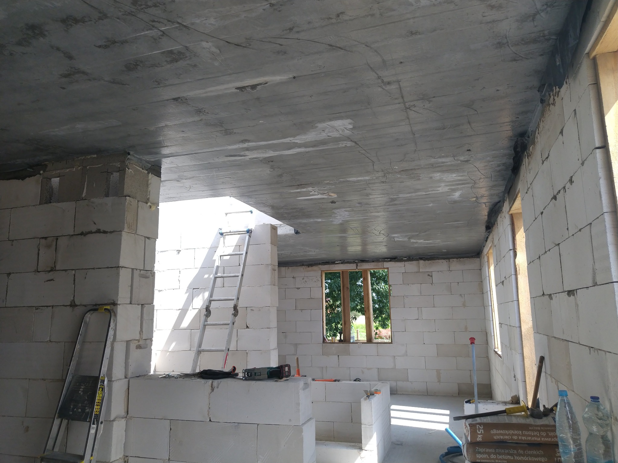 Wnętrze domu w trakcie budowy, widoczne ściany z betonu komórkowego, surowy betonowy strop, drabina oparta o ścianę, narzędzia budowlane i materiały, okno z widokiem na zieleń.