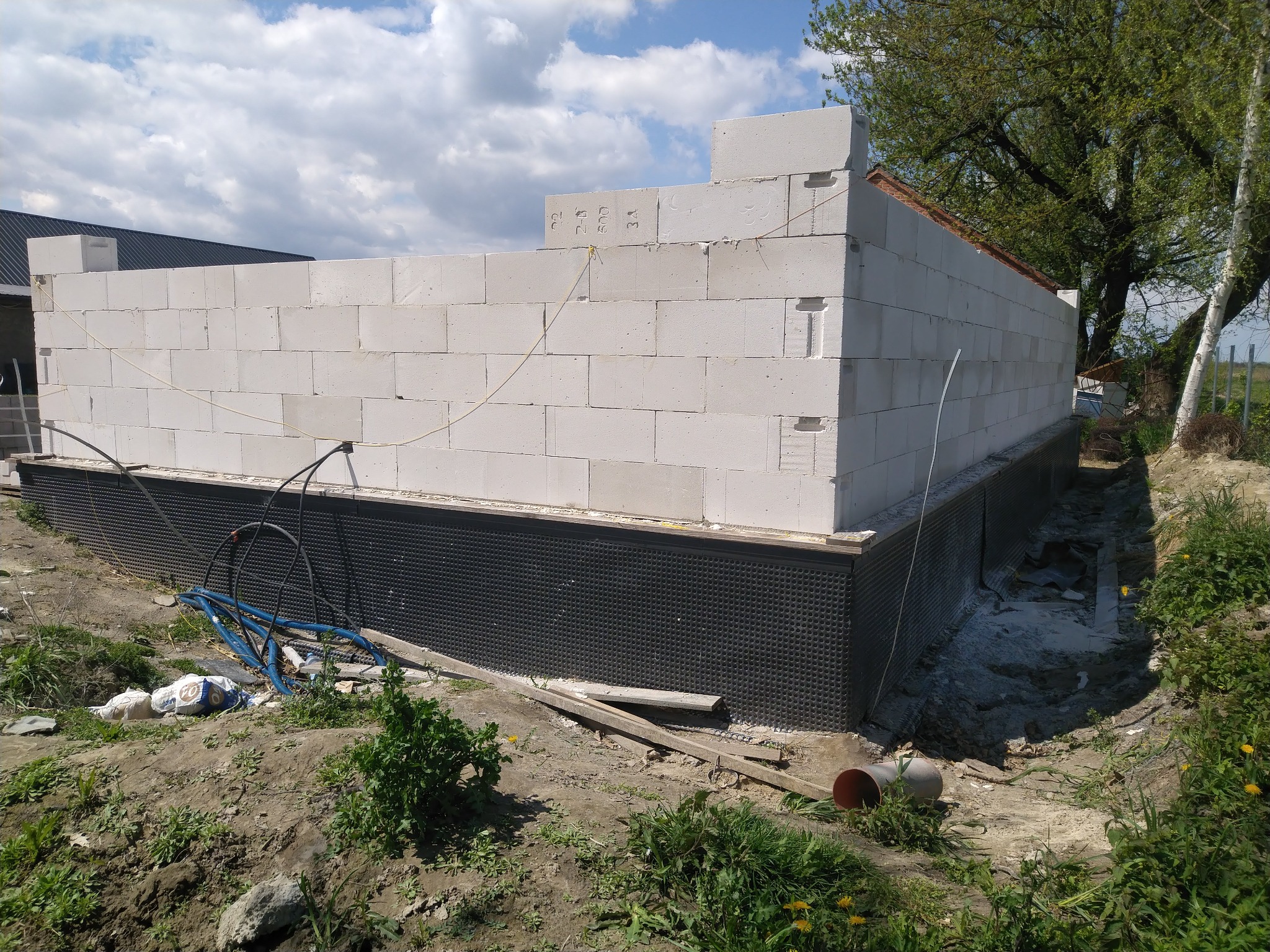 Wznoszone ściany parteru z betonu komórkowego na fundamencie z hydroizolacją, widoczne worki z zaprawą i niebieskie rury na placu budowy, w tle drzewa i błękitne niebo z chmurami.