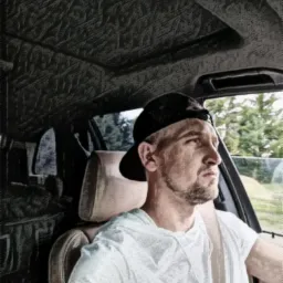 Mężczyzna w czapce bejsbolówce siedzi w samochodzie, patrzy w prawo. Widoczne wnętrze auta i drzewa za oknem. Stylizacja zdjęcia przypomina szkic.