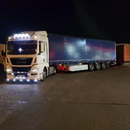 Biała ciężar&oacute;wka MAN EfficientLine z naczepą pod plandeką, oświetlona reflektorami na dachu, zaparkowana nocą na brukowanym placu.