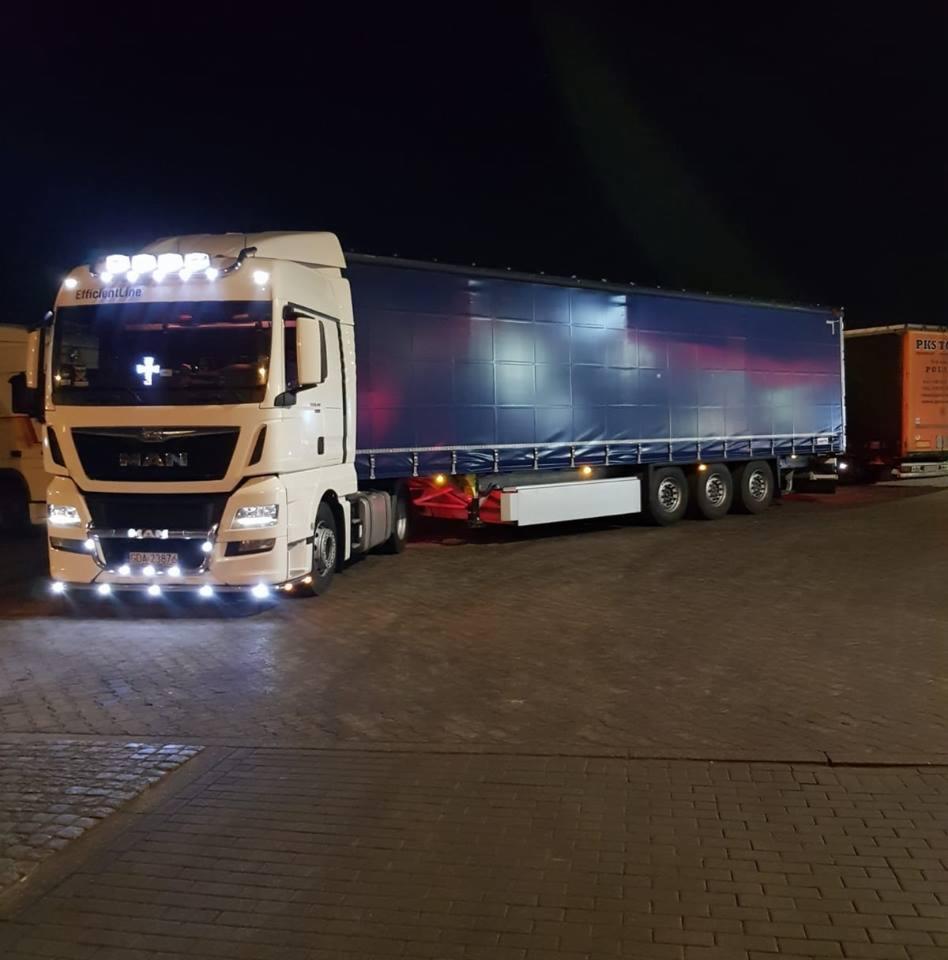 Biała ciężarówka MAN EfficientLine z naczepą pod plandeką, oświetlona reflektorami na dachu, zaparkowana nocą na brukowanym placu.