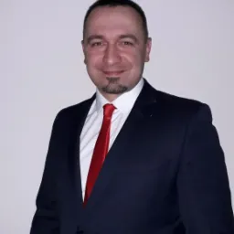 Portret mężczyzny w ciemnym garniturze i czerwonym krawacie, z delikatnym uśmiechem i kozia bródką, na jasnym, jednolitym tle.