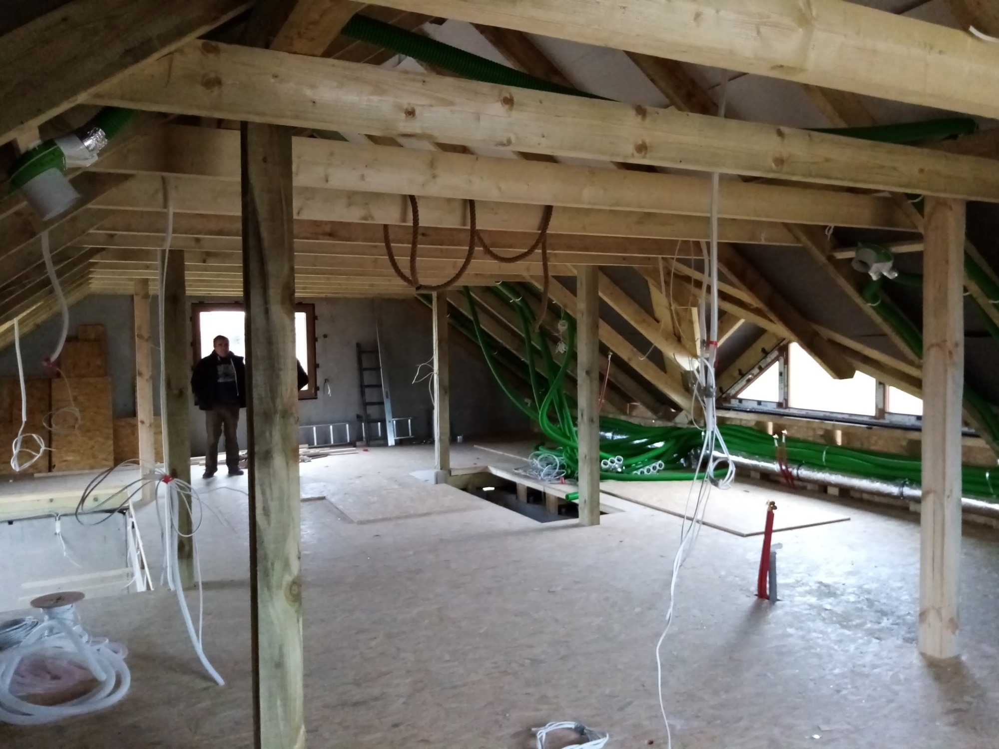 Poddasze w trakcie adaptacji: widoczne drewniane belki konstrukcyjne, instalacja wentylacyjna z zielonych rur, elektryka w trakcie montażu, osoba w oknie.