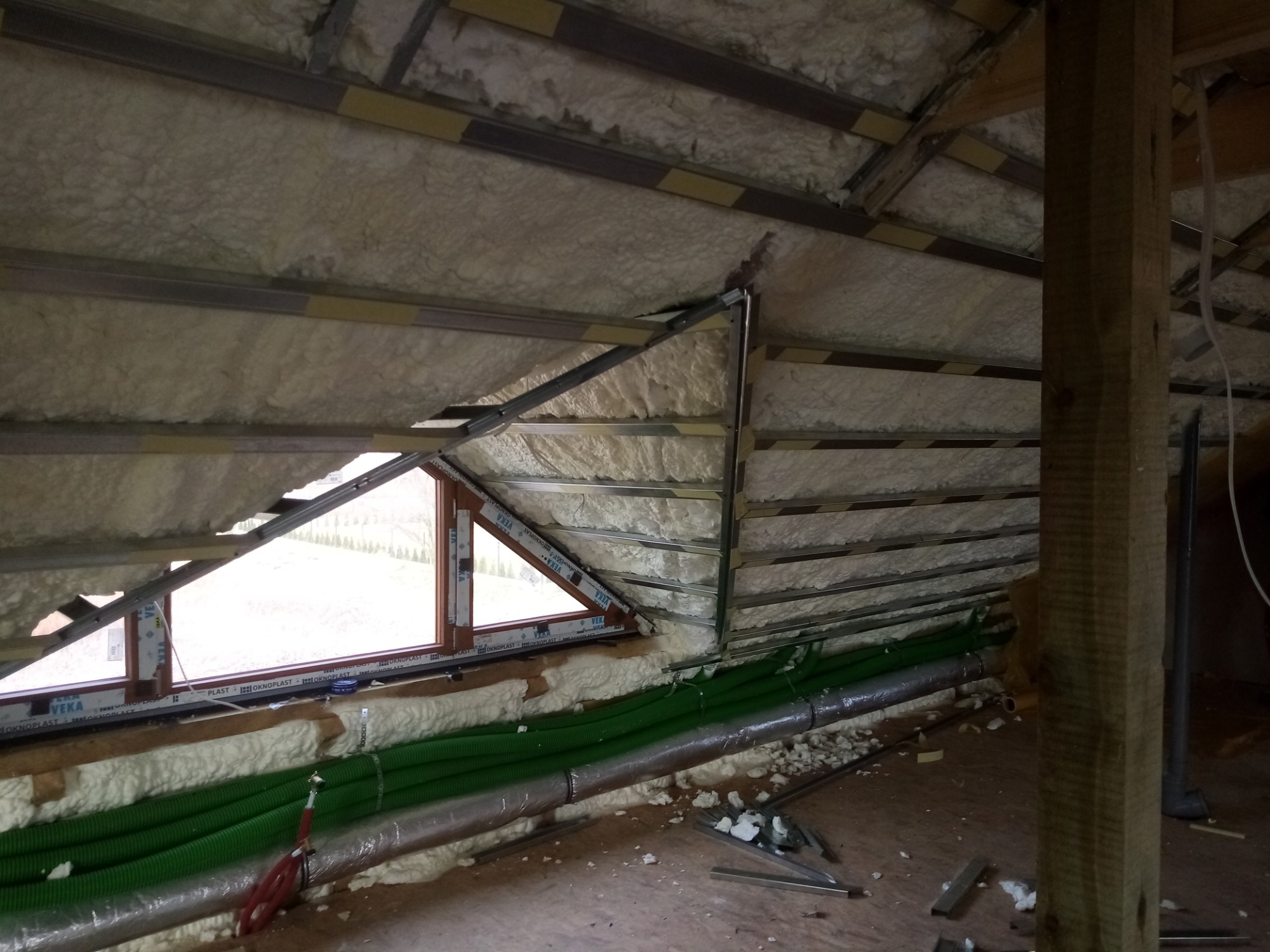 Ocieplenie poddasza pianką PUR, widoczne metalowe profile, okno trójkątne i zielone rury wentylacyjne w trakcie prac.