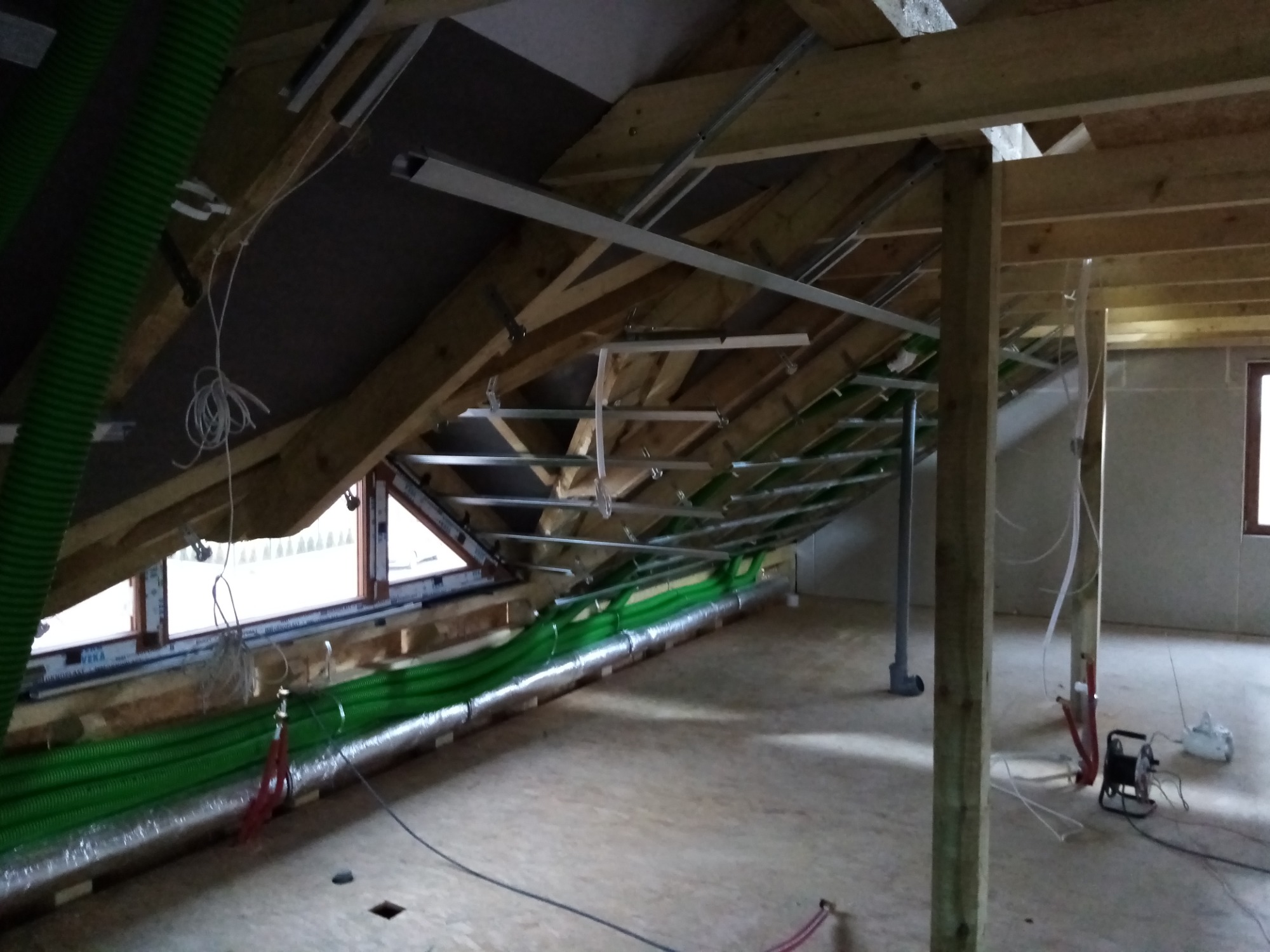 Poddasze w trakcie adaptacji: widoczne ocieplenie, konstrukcja drewniana dachu, instalacja wentylacyjna z zielonymi rurami, metalowe profile pod płyty gipsowo-kartonowe, okno dachowe w drewnianej...