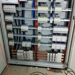 Krzysztof Wojciulewicz PROEL - Otwarta szafa elektryczna z widocznymi zabezpieczeniami, przewodami i listwami. Na podłodze narzędzia: wiertarka, śrubokręty, zwój kabla i dokumentacja.