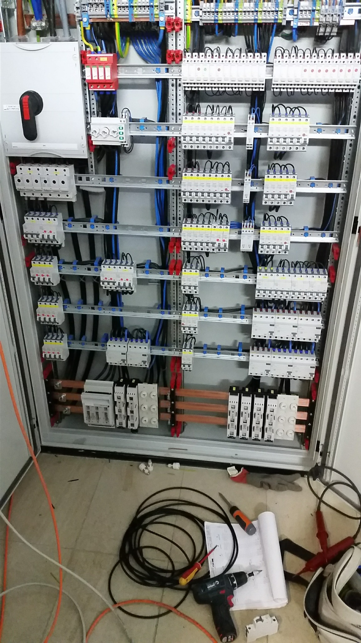 Otwarta szafa elektryczna z widocznymi zabezpieczeniami, przewodami i listwami. Na podłodze narzędzia: wiertarka, śrubokręty, zwój kabla i dokumentacja.