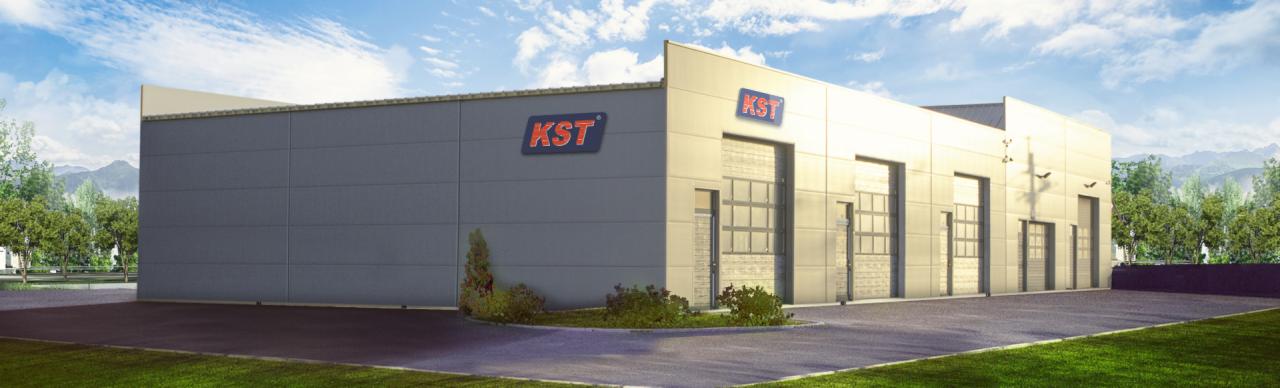 Nowoczesny budynek przemysłowy z szarymi ścianami panelowymi, przeszklonymi bramami segmentowymi i logo firmy KST na elewacji, otoczony zielenią i błękitnym niebem z chmurami.