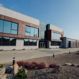 Nowoczesny budynek biurowy z dużymi oknami i elementami z blachy w kolorze rdzy, otoczony zagospodarowanym terenem z kamieniami i roślinami, z widoczną flagą firmy w tle.