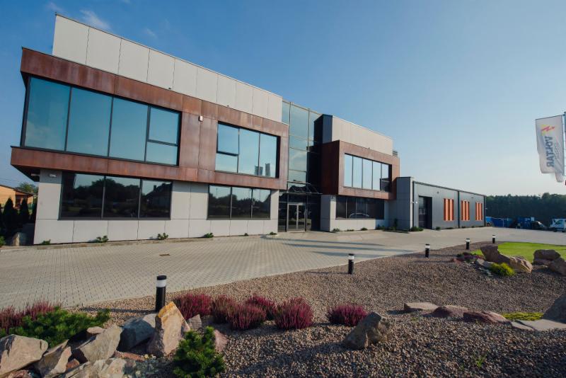 Nowoczesny budynek biurowy z dużymi oknami i elementami z blachy w kolorze rdzy, otoczony zagospodarowanym terenem z kamieniami i roślinami, z widoczną flagą firmy w tle.