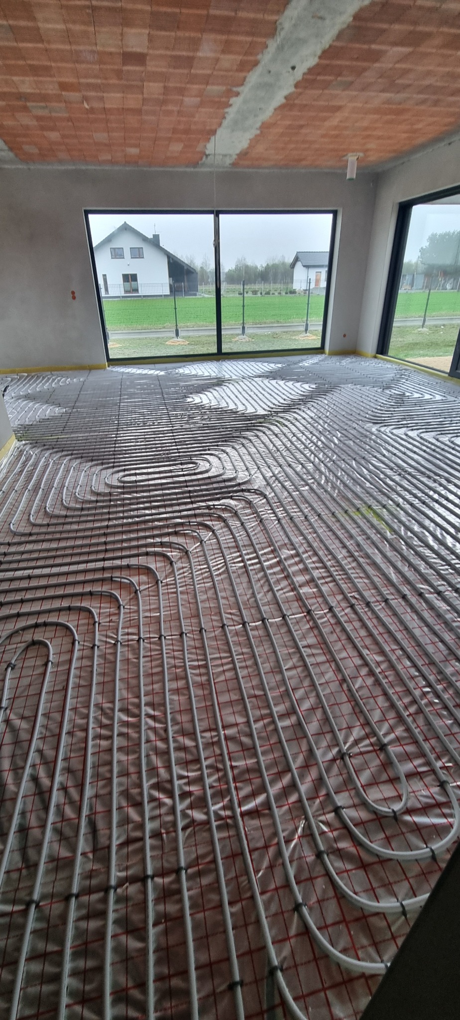 Szare rury ogrzewania podłogowego ułożone na folii z czerwoną siatką w surowym wnętrzu. W tle okno z widokiem na dom i zielony trawnik.