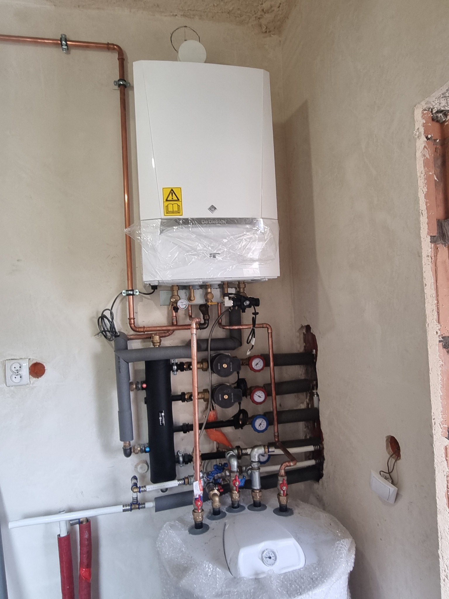 Nowy, biały piec gazowy De Dietrich, jeszcze owinięty folią ochronną, z podłączonymi miedzianymi rurami i czarnymi izolacjami, zamontowany na szarej ścianie w surowym stanie.