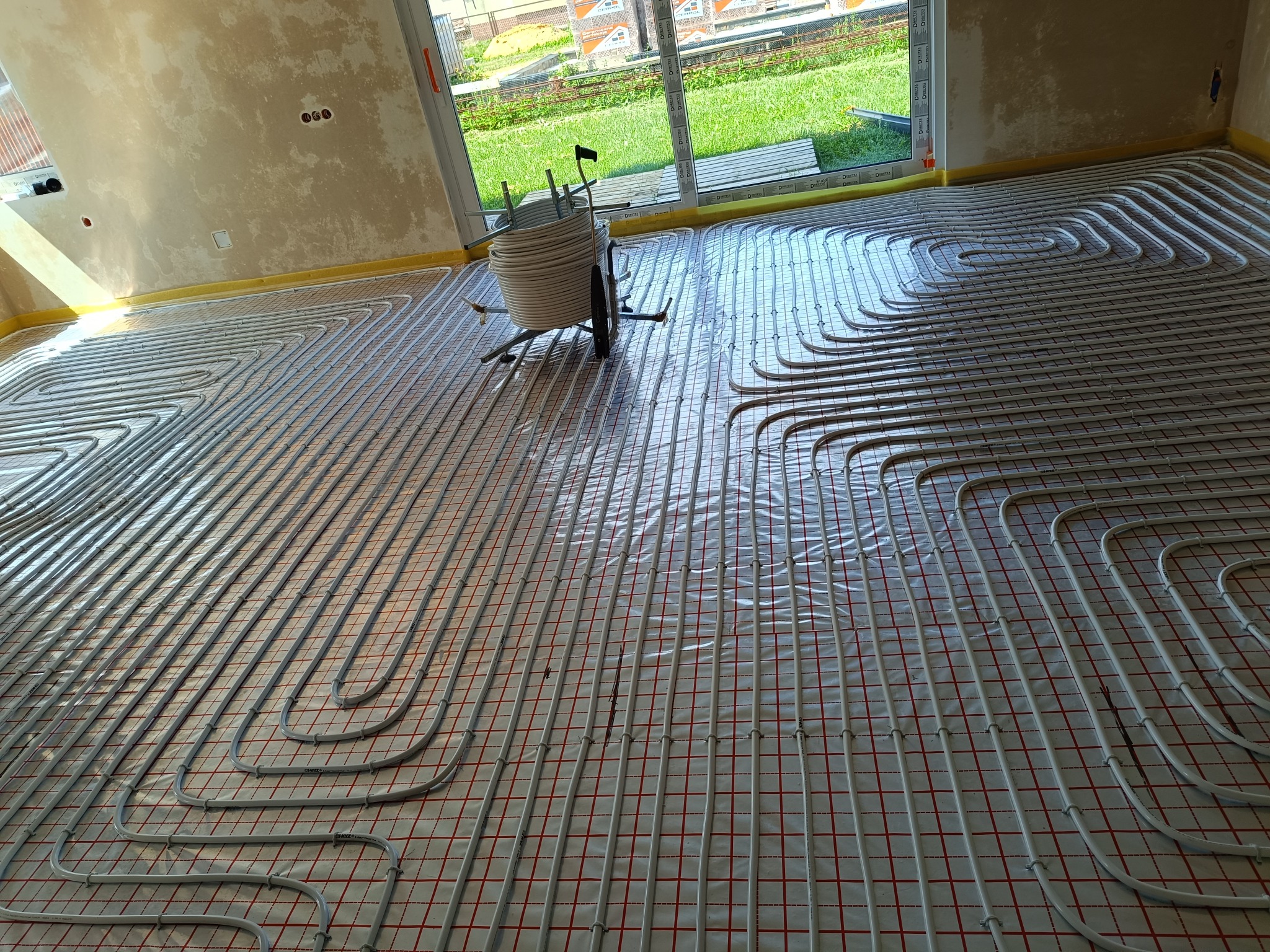 Białe rury ogrzewania podłogowego ułożone na czerwonej siatce w jasnym pomieszczeniu z oknem. Widoczny wózek z rolką rur. Etap instalacji przed wylaniem posadzki.