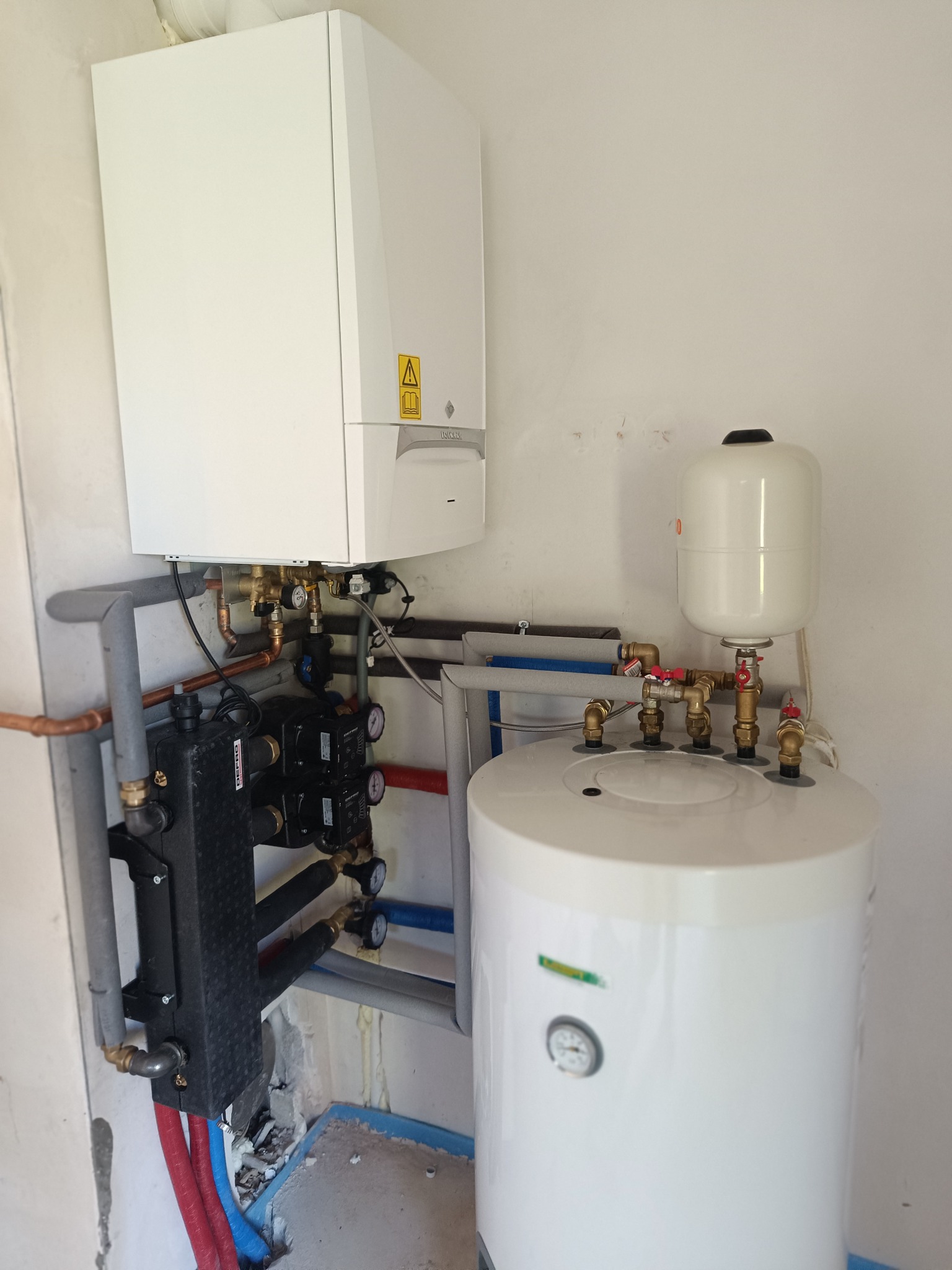 Biały piec gazowy kondensacyjny z zasobnikiem i osprzętem, widoczne rury miedziane i manometry, zainstalowany na ścianie.