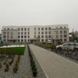 Nowoczesny blok mieszkalny z jasną elewacją, otoczony zagospodarowanym terenem zielonym z chodnikiem z kostki i ozdobnymi krzewami, samoch&oacute;d osobowy zaparkowany w pobliżu.