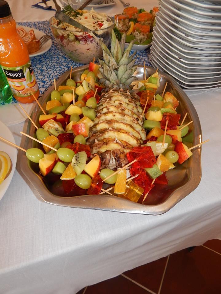 Srebrna patera z ananasem, owocami (winogrona, kiwi, pomarańcze, jabłka) i galaretką na szpadkach, uzupełniona sałatką, roladkami i napojem Cappy w tle.