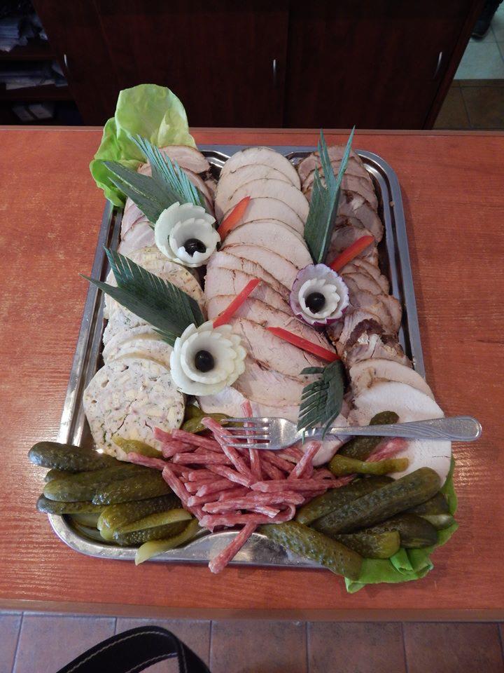 Srebrna taca z wędlinami w plasterkach, dekoracjami z warzyw i kwiatów z oliwek, uzupełniona o korniszony i cienkie kiełbaski, z widocznym widelcem.