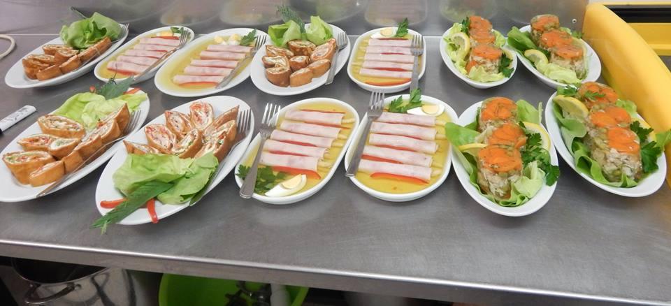 Różnorodne przystawki na białych owalnych talerzach: roladki z ciasta, szynka w galarecie z jajkiem i papryką, sałatka warstwowa z marchewką, podane na metalowym blacie.