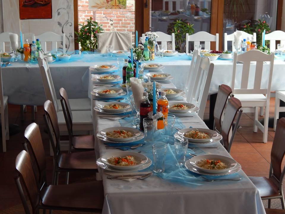 Długi stół nakryty do posiłku, białe obrusy, niebieski tiul, białe i brązowe krzesła, na talerzach danie z makaronu i warzyw, napoje w butelkach, kwiaty w wazonach, widok przez okno na ceglany mur.