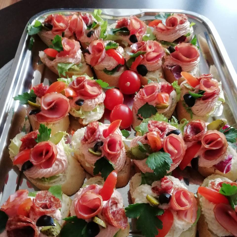 Srebrna taca z apetycznymi kanapeczkami: pieczywo z kremowym smarowidłem, dekorowane rożkami szynki, warzywami i oliwkami; trzy pomidorki koktajlowe w centrum.