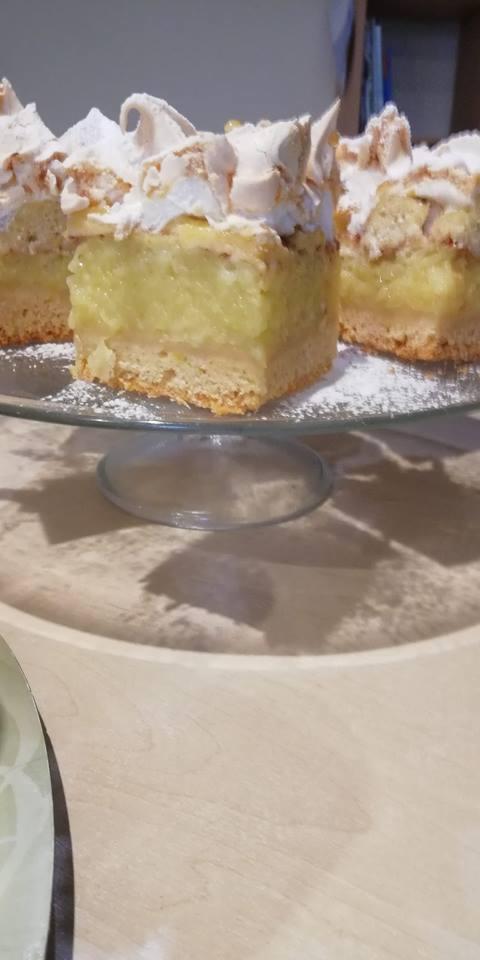 Kawałki domowego ciasta z jabłkami i bezową pianką na szklanym stojaku, posypane cukrem pudrem.