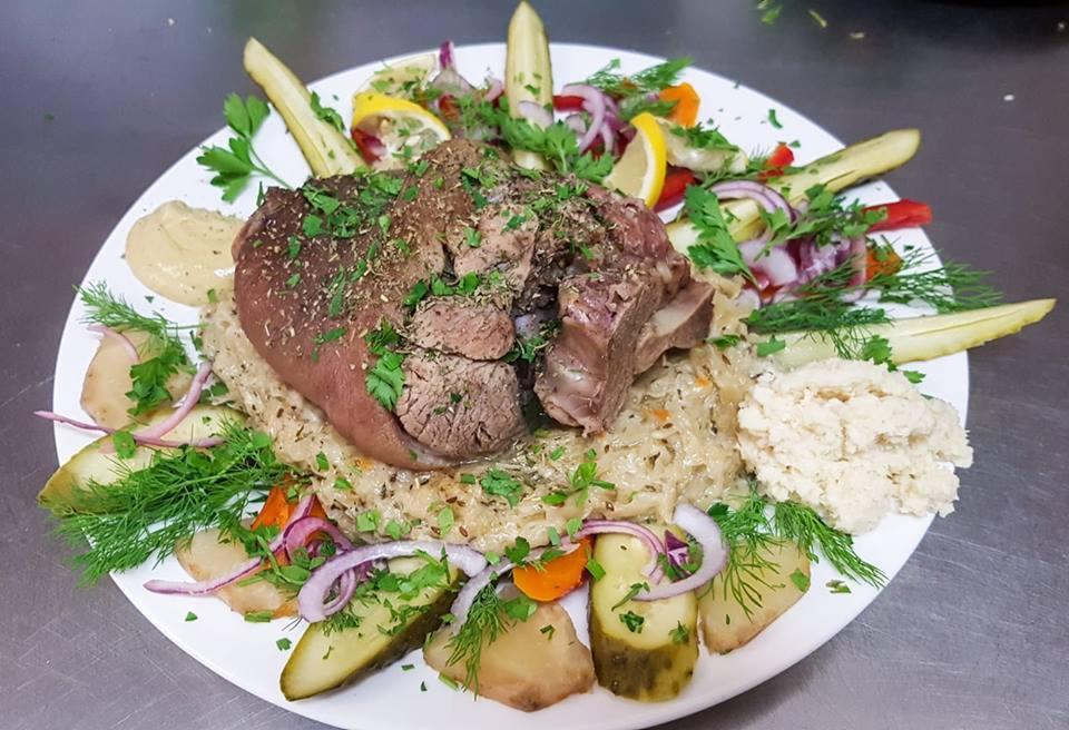 Pieczona golonka wieprzowa z kapustą, ziemniakami, ogórkiem kiszonym, czerwoną cebulą, cytryną i sosem na białym talerzu.