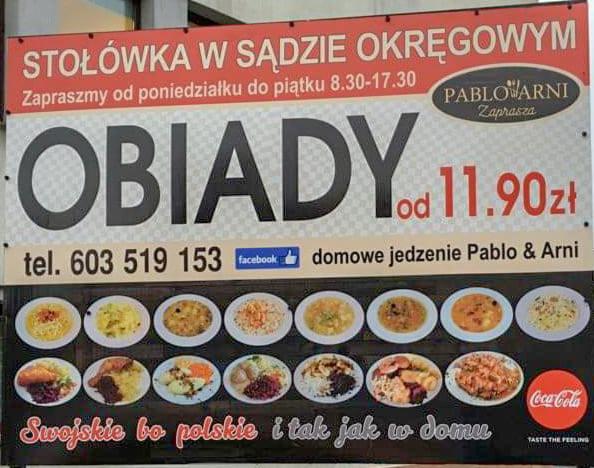 Szyld stołówki w sądzie okręgowym z ofertą obiadów domowych Pablo & Arni, prezentujący zdjęcia dań i logo Coca-Coli.