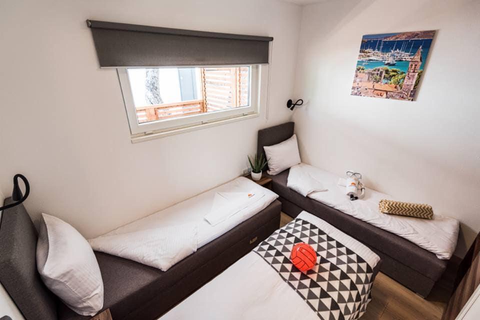 Wnętrze pokoju z trzema łóżkami, z widokiem na okno z roletą i obrazem portu morskiego na ścianie.