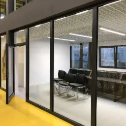 Przeszklona sala konferencyjna z czarnymi ramami okiennymi i drzwiowymi, żółta podłoga, widoczne krzesła i biurka wewnątrz, otwarte drzwi z klamką po lewej stronie.