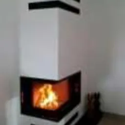 Biały narożny piec z czarnymi akcentami dekoracyjnymi na obudowie, płomienie widoczne przez przeszklone drzwiczki, czarna podstawa pieca, minimalistyczny design.