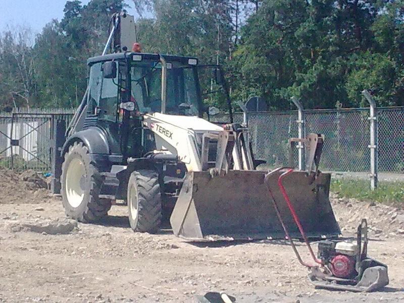 Koparko-ładowarka Terex na placu budowy, obok ubijak wibracyjny, w tle ogrodzenie z siatki i drzewa.