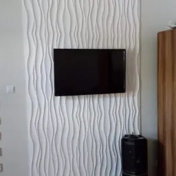 Biała, trójwymiarowa panelowa ściana z falistym wzorem, na której zawieszony jest czarny telewizor. Obok stoi drewniana szafka i czarny głośnik.