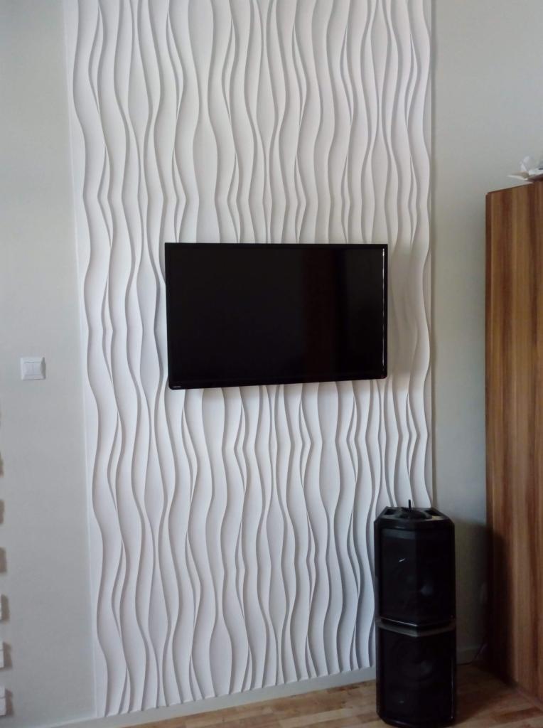 Biała, trójwymiarowa panelowa ściana z falistym wzorem, na której zawieszony jest czarny telewizor. Obok stoi drewniana szafka i czarny głośnik.