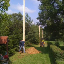 Montaż drewnianych słupów w ogrodzie z użyciem koparki, dwóch mężczyzn stabilizuje pierwszy słup, widoczna sterta ziemi.