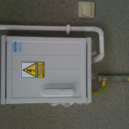 Szara skrzynka elektryczna Pelmet z ostrzeżeniem przed porażeniem prądem, zamontowana na szarej, chropowatej ścianie. Nad skrzynką biała rura PCV, z boku widoczny przewód uziemiający i listwa.