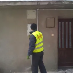 Mężczyzna w odblaskowej kamizelce firmy FORBUD-2 sprawdza skrzynkę z bezpiecznikami na elewacji budynku, widoczny fragment rury instalacji elektrycznej i drewniane drzwi wejściowe.