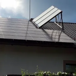 Panele słoneczne na dachu domu jednorodzinnego z dodatkowym panelem zamontowanym na stelażu, widok z dołu na tle pochmurnego nieba.